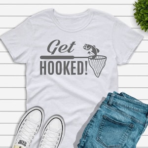May include: White t-shirt with the text "Get Hooked!" and a graphic of a fish being caught in a net. The shirt is laid out on a white wooden surface with a pair of white sneakers and blue jeans.
