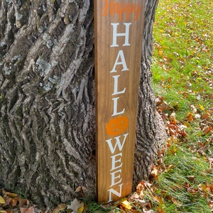 Pode incluir: Uma placa de madeira com as palavras "Happy Halloween" escritas em tinta branca. A placa &eacute; decorada com um morcego preto e uma ab&oacute;bora laranja.