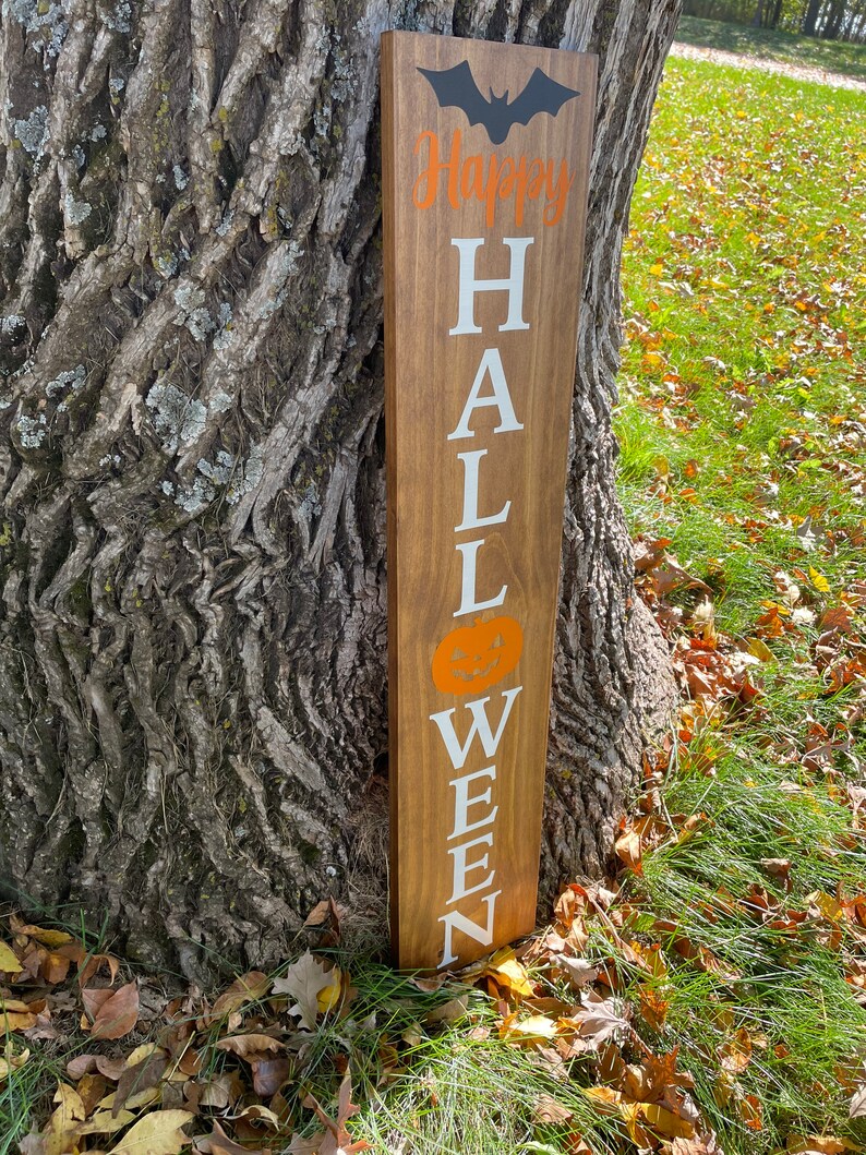 Pode incluir: Uma placa de madeira com as palavras "Happy Halloween" escritas em tinta branca. A placa &eacute; decorada com um morcego preto e uma ab&oacute;bora laranja.