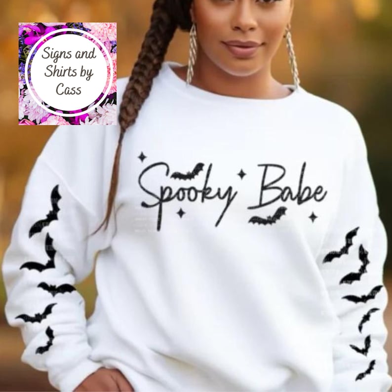以下が含まれることがあります： 「Spooky Babe」の文字が黒い筆記体で書かれた白いスウェットシャツ。コウモリのシルエットと星で装飾されています。袖にはコウモリのデザインがあります。カジュアルなハロウィーンをテーマにしたアパレルアイテムです。