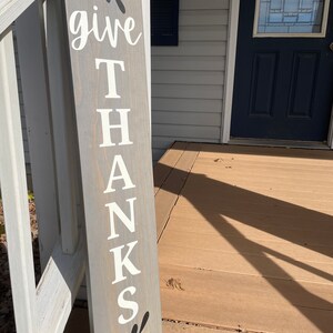 Pode incluir: Uma placa de madeira cinza com as palavras "give thanks" escritas em tinta branca. A placa tem um visual r&uacute;stico e &eacute; decorada com desenhos de folhas pretas.