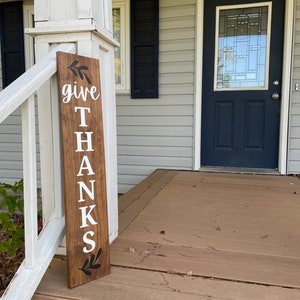 Pode incluir: Uma placa de madeira com as palavras "give thanks" pintadas de branco sobre um fundo castanho. A placa &eacute; decorada com folhas pretas na parte superior e inferior.