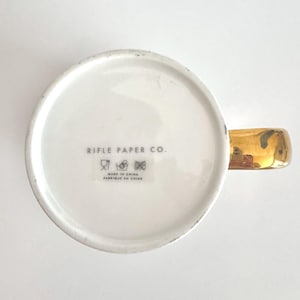 May include: White ceramic mug with a gold-colored handle. The bottom of the mug features the text "RIFLE PAPER CO." and "MADE IN CHINA FABRIQUE EN CHINE". The mug has a simple, elegant design.