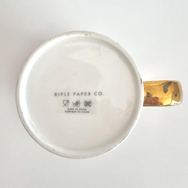 May include: White ceramic mug with a gold-colored handle. The bottom of the mug features the text "RIFLE PAPER CO." and "MADE IN CHINA FABRIQUE EN CHINE". The mug has a simple, elegant design.