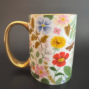 May include: White ceramic mug with a gold handle and rim, adorned with a colorful floral pattern. The design includes red, yellow, pink, and blue flowers, along with green leaves and a brown butterfly.