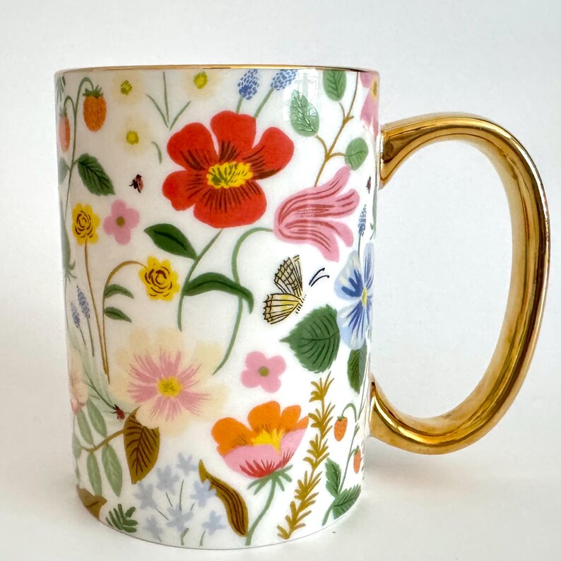 May include: White ceramic mug with a colorful floral design featuring red, pink, yellow, and blue flowers, strawberries, and a butterfly. The mug has a gold-colored handle.