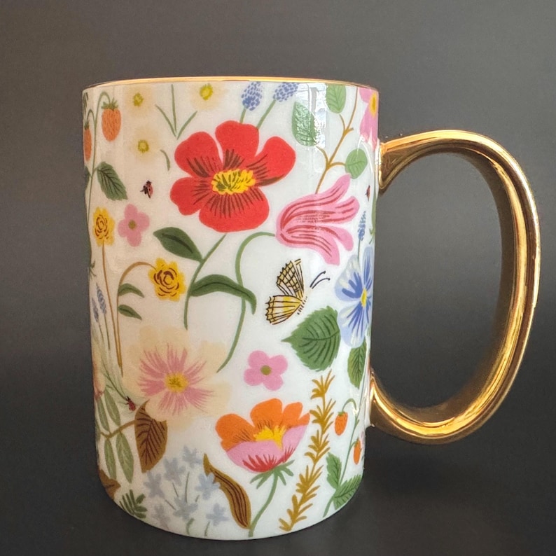 May include: White ceramic mug with a gold handle and rim, adorned with a colorful floral pattern. The design includes red, pink, orange, and blue flowers, along with butterflies and green leaves. A decorative mug for beverages.