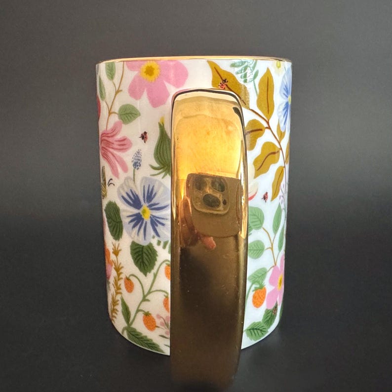 May include: A white ceramic mug with a gold handle and rim, adorned with a floral pattern of pink, blue, and yellow flowers, green leaves, and small red berries. The mug has a glossy finish.