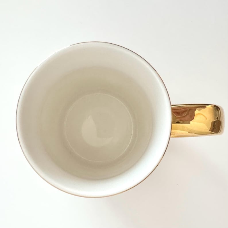 May include: A white ceramic mug with a gold-colored handle and rim. The interior of the mug is white, and the handle reflects the light. The mug is set against a white background.