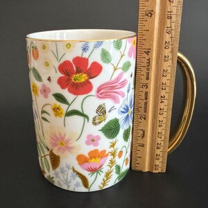 May include: White ceramic mug with a gold handle and rim, adorned with a colorful floral pattern. The design includes red, pink, orange, and blue flowers, green leaves, and a yellow butterfly. The mug is approximately 4 inches tall.