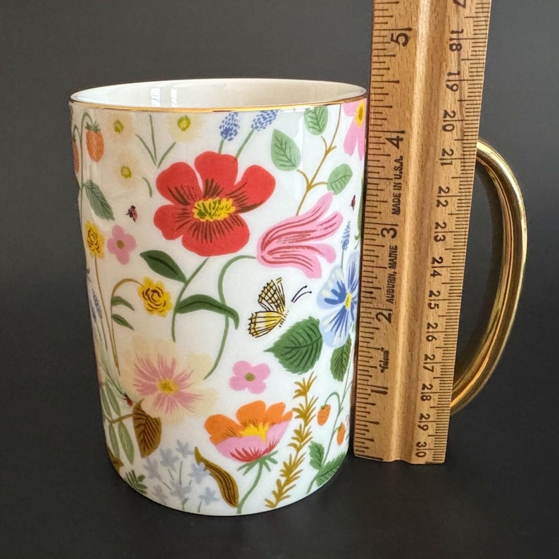 May include: White ceramic mug with a gold handle and rim, adorned with a colorful floral pattern. The design includes red, pink, orange, and blue flowers, green leaves, and a yellow butterfly. The mug is approximately 4 inches tall.