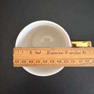 May include: A white ceramic teacup with a gold handle, measured with a wooden ruler. The ruler shows the cup is approximately 4 inches in diameter. The cup is on a black surface.