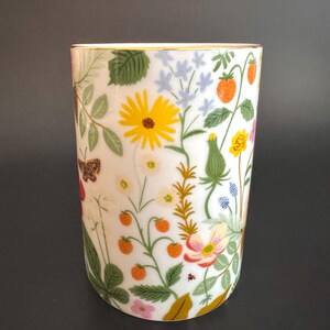 May include: A white ceramic tumbler with a gold rim, adorned with a colorful floral design. The pattern includes yellow sunflowers, orange strawberries, and various wildflowers, along with green leaves and a butterfly. The tumbler has a cylindrical shape.
