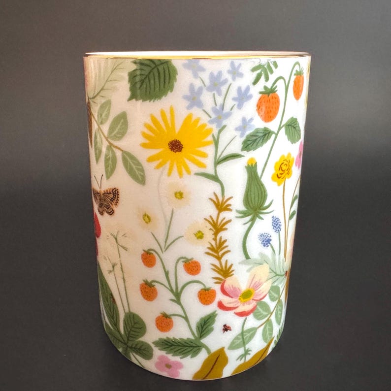 May include: A white ceramic tumbler with a gold rim, adorned with a colorful floral design. The pattern includes yellow sunflowers, orange strawberries, and various wildflowers, along with green leaves and a butterfly. The tumbler has a cylindrical shape.