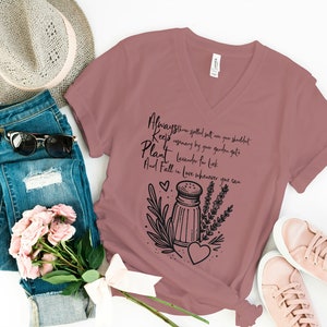 K&ouml;nnte beinhalten: Ein mauvefarbenes T-Shirt mit V-Ausschnitt und einem schwarzen Grafikdesign mit einem Salzstreuer, Lavendel und einem Herzen. Der Text auf dem Shirt lautet "Always throw spilled salt on your shoulder, Keep rosemary by your garden gate, Plant lavender for luck, And Fall in love whenever you can."