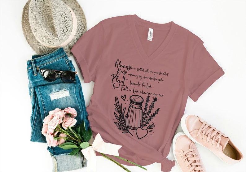 K&ouml;nnte beinhalten: Ein mauvefarbenes T-Shirt mit V-Ausschnitt und einem schwarzen Grafikdesign mit einem Salzstreuer, Lavendel und einem Herzen. Der Text auf dem Shirt lautet "Always throw spilled salt on your shoulder, Keep rosemary by your garden gate, Plant lavender for luck, And Fall in love whenever you can."