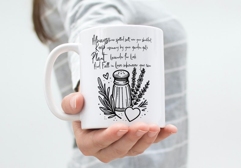 K&ouml;nnte beinhalten: Eine wei&szlig;e Keramiktasse mit einer schwarz-wei&szlig;en Illustration eines Salzstreuers, Rosmarins und Lavendels. Der Text auf der Tasse lautet "Always store spilled salt over your shoulder Keep rosemary by your garden gate Plant lavender for luck And Fall in love whenever you can".