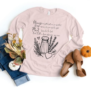 May include: A pink long-sleeved shirt with a black graphic design featuring a salt shaker, herbs, and a heart. The text on the shirt reads "Always throw spelled salt over your shoulder Keep rosemary by your garden gate Plant lavender for luck And Fall in love whenever you can."