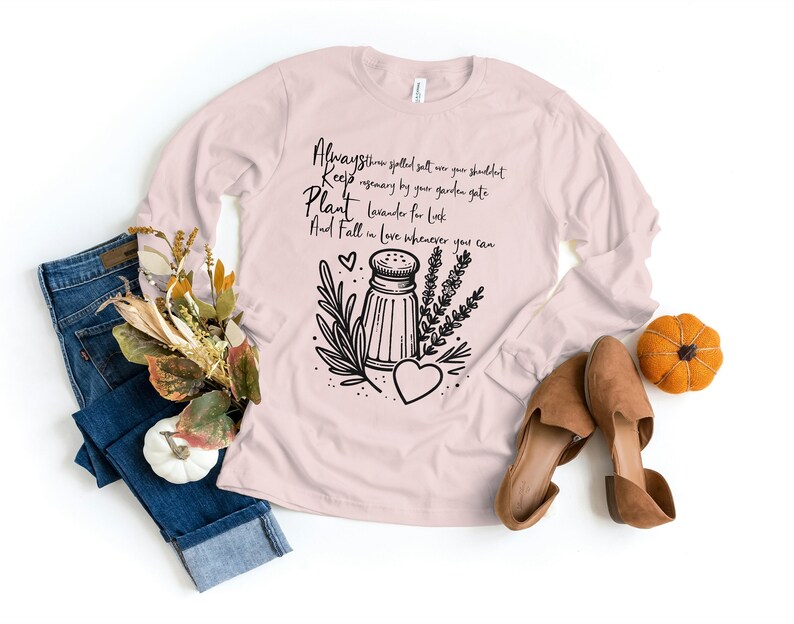 K&ouml;nnte beinhalten: Ein rosa Langarmshirt mit einem schwarzen grafischen Design, das einen Salzstreuer, Kr&auml;uter und ein Herz zeigt. Der Text auf dem Shirt lautet "Always throw spelled salt over your shoulder Keep rosemary by your garden gate Plant lavender for luck And Fall in love whenever you can."