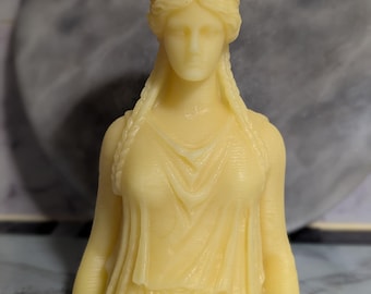 Puede incluir: Una vela amarilla con forma de busto de una mujer que lleva una corona. La vela tiene un rostro y un cabello detallados.