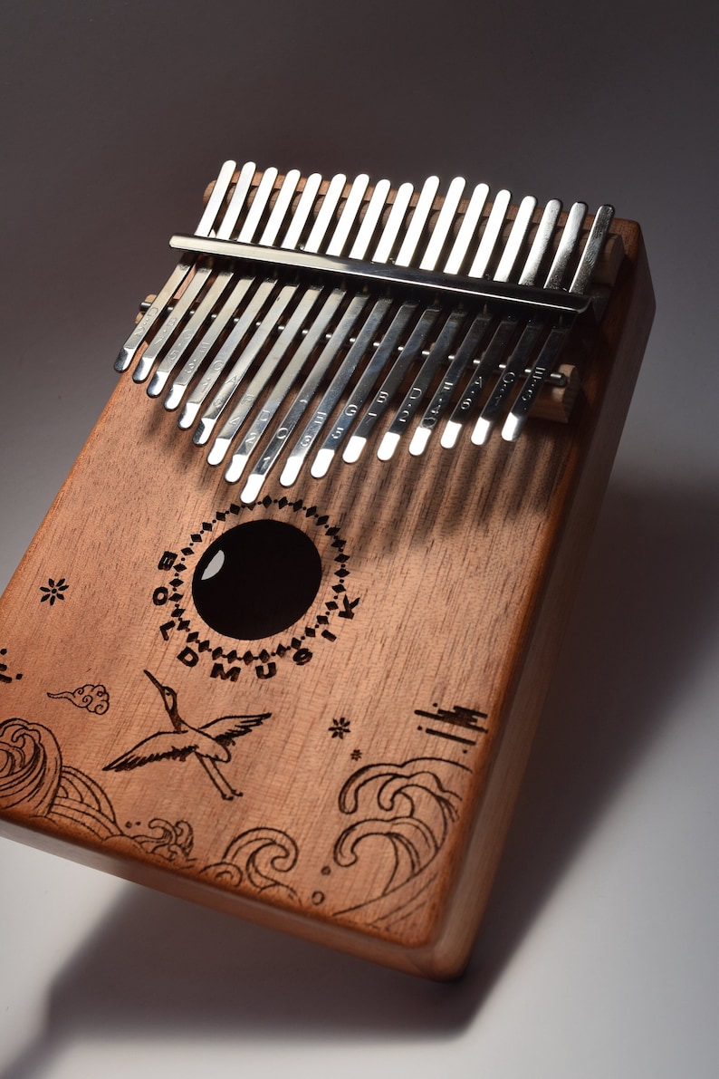 Main image of a kalimba thumb piano 17 key in studio lighting. It is Bold Musik brand, and itis an open hole design with a bird and ocean waves carved into the mahogany wood box frame. The tines or keys are made of stainless steel.