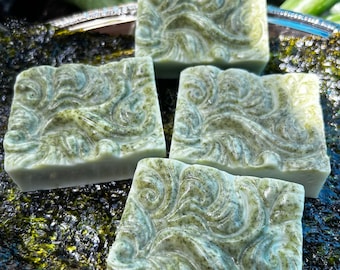 May include: Four square bars of green soap with a swirling pattern. The soap is sitting on a bed of dark green moss.