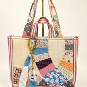 Pode incluir: Uma bolsa tote acolchoada feita de vários retalhos de tecido com padrões florais, xadrez e sólidos. A bolsa apresenta painéis laterais e alças com listras vermelhas e brancas, com um pingente decorativo preso à alça frontal com tiras de tecido azul.