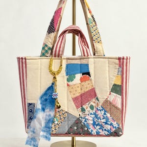Pode incluir: Uma bolsa tote acolchoada feita de vários retalhos de tecido com padrões azuis, vermelhos, amarelos e brancos. A bolsa apresenta painéis laterais e alças com listras vermelhas e brancas, e um chaveiro de metal dourado com uma tira de tecido azul tie-dye.