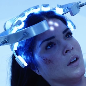 May include: A woman wearing a futuristic head device with glowing blue lights. The device is metallic and has a series of lights across the forehead and around the head. The woman is looking up with a concerned expression.