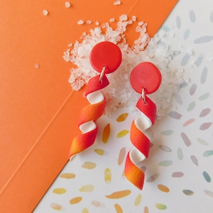 Puede incluir: Un par de pendientes de arcilla polimérica de color rojo y naranja con detalles blancos. Los pendientes cuelgan de tapas de disco rojo.