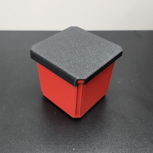 May include: A small, square container with a red body and a textured, dark gray lid. The lid has a black trim. The container is sitting on a black surface.
