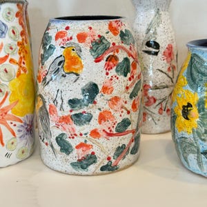 May include: A collection of hand-painted ceramic vases. The vases feature colorful floral and bird designs in shades of orange, yellow, blue, and green against a speckled white background. The vases vary in shape and size.