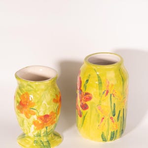 May include: Two ceramic vases with floral designs. The vases are painted with a bright yellow background and feature orange and red flowers with green stems and leaves. The vase on the left is shorter and wider, while the one on the right is taller and cylindrical.