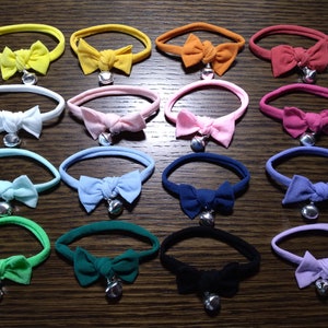 May include: A collection of colorful cat collars with bowties and silver bell charms. The collars are in various colors, including yellow, orange, red, pink, blue, green, and black. Each collar has a small, shiny bell attached.