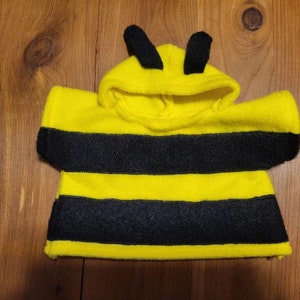 Puede incluir: Un disfraz de abeja amarillo y negro para niño. El disfraz es de fieltro y presenta una capucha con antenas negras. El cuerpo del disfraz tiene rayas negras horizontales. Diseñado para ser usado sobre la ropa del niño.