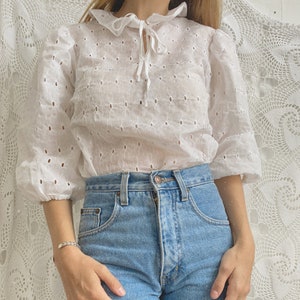 Puede incluir: Una blusa blanca de encaje con cuello de volantes y un lazo en el cuello. La blusa tiene un corte holgado y mangas 3/4. La modelo lleva la blusa con jeans azules.