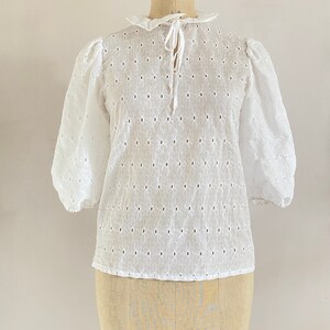 Puede incluir: Blusa blanca de algod&oacute;n con un delicado estampado floral y un lazo en el cuello.