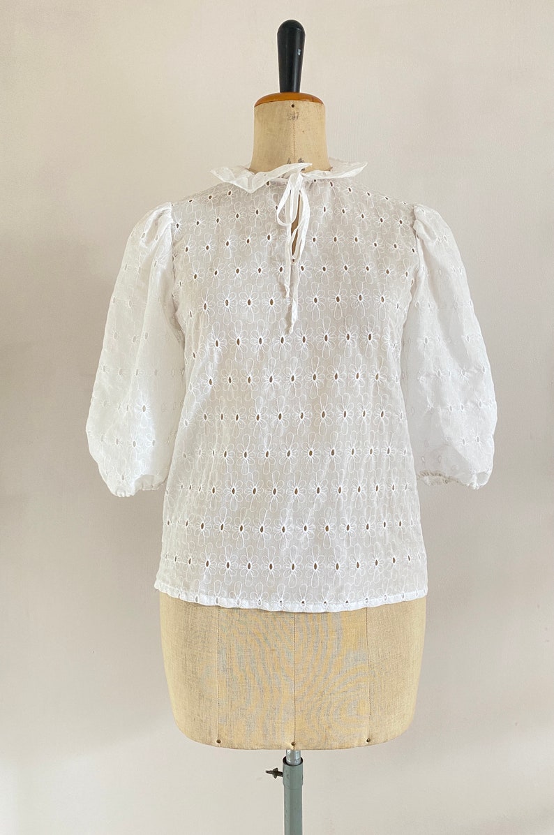 Puede incluir: Blusa blanca de algod&oacute;n con un delicado estampado floral y un lazo en el cuello.