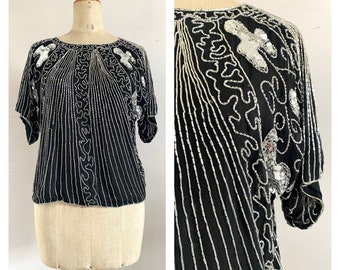 Puede incluir: Una blusa negra con lentejuelas y bordados plateados y un escote redondo. La blusa tiene mangas cortas y un corte holgado.