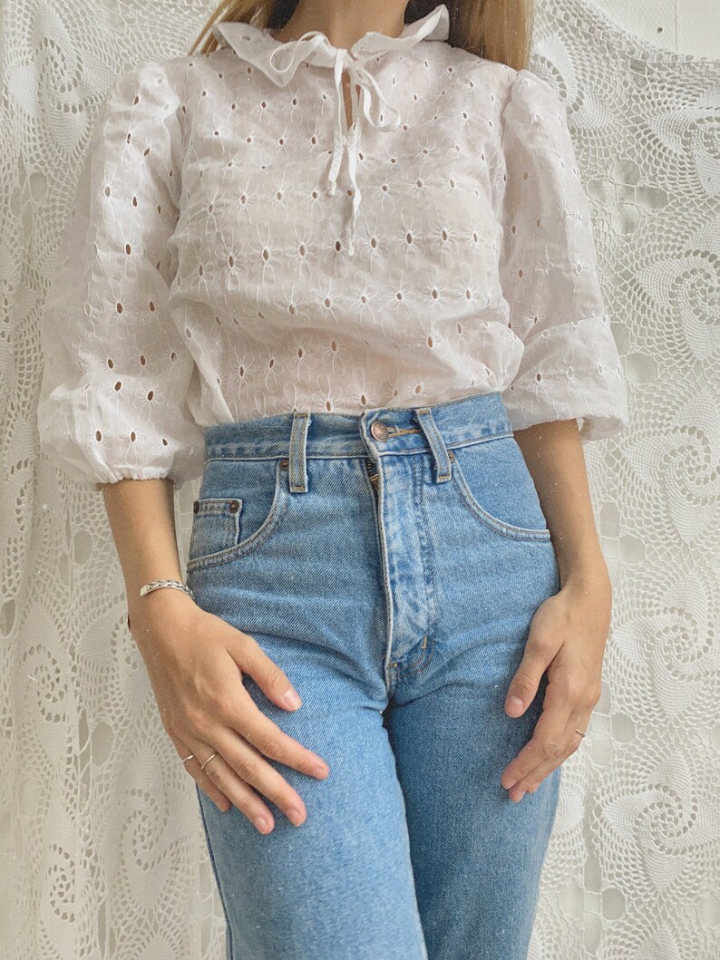 Puede incluir: Una blusa blanca de encaje con cuello de volantes y un lazo en el cuello. La blusa tiene un corte holgado y mangas 3/4. La modelo lleva la blusa con jeans azules.