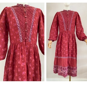 Könnte beinhalten: Ein langärmeliges, rotes Blumenkleid mit gestepptem Oberteil und Knopfleiste. Das Kleid hat einen hohen Ausschnitt und einen gerafften Rock.