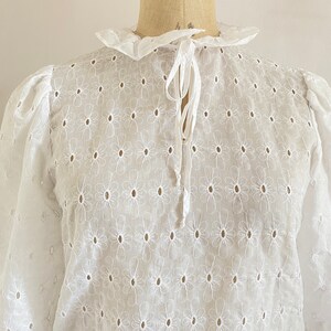 Puede incluir: Blusa blanca de algod&oacute;n con un delicado patr&oacute;n de bordado floral y un lazo en el cuello.