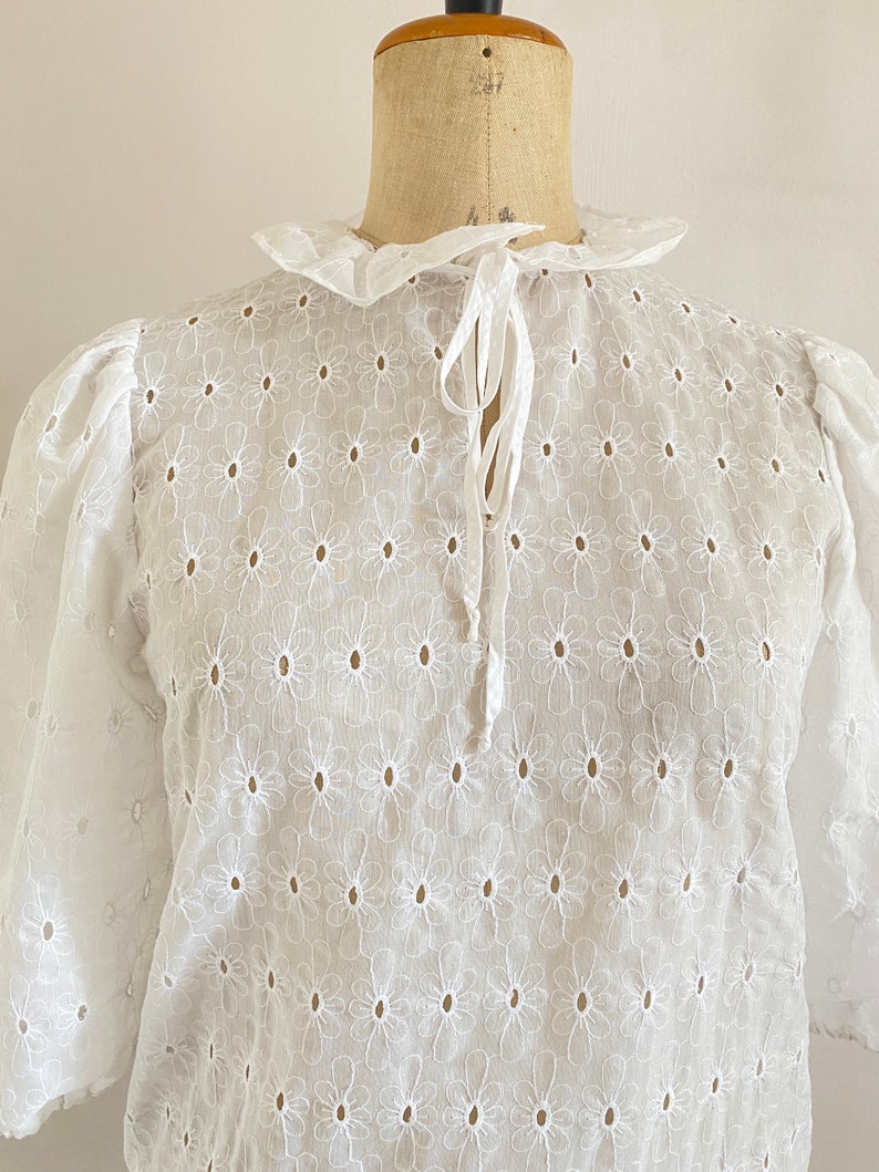 Puede incluir: Blusa blanca de algod&oacute;n con un delicado patr&oacute;n de bordado floral y un lazo en el cuello.