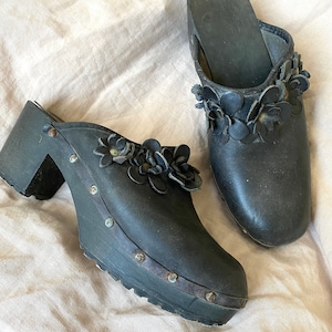 May include: A pair of black leather clogs with flower embellishments. The clogs have a wooden platform and a chunky heel.