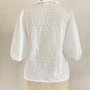 Puede incluir: Blusa blanca de algod&oacute;n con un delicado patr&oacute;n de ojal floral y manga abullonada. La blusa tiene un escote redondo y un cierre de bot&oacute;n en la espalda.