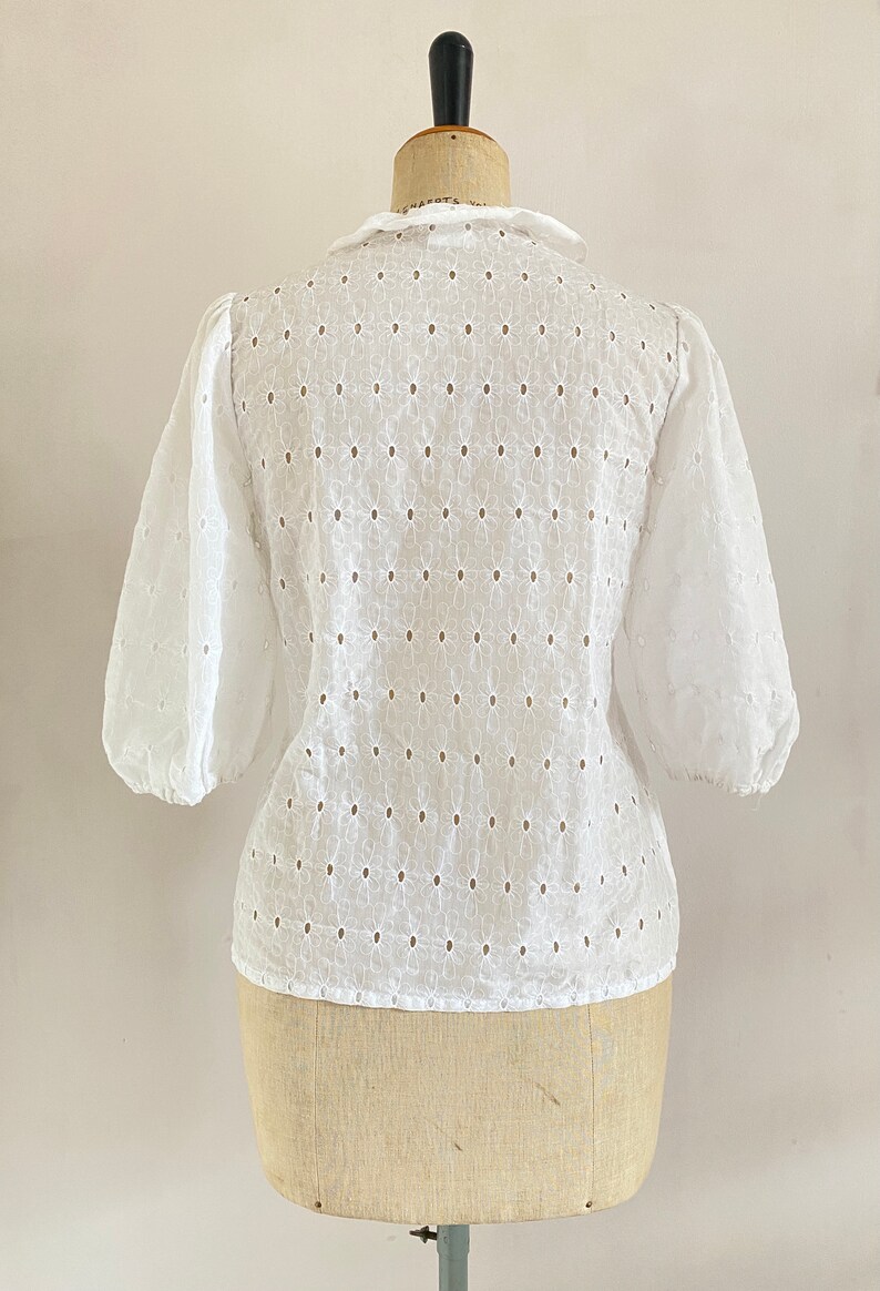 Puede incluir: Blusa blanca de algod&oacute;n con un delicado patr&oacute;n de ojal floral y manga abullonada. La blusa tiene un escote redondo y un cierre de bot&oacute;n en la espalda.