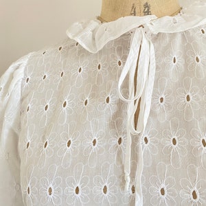Puede incluir: Blusa blanca de algod&oacute;n con cuello fruncido y un lazo en el cuello. La blusa est&aacute; bordada con un patr&oacute;n floral.