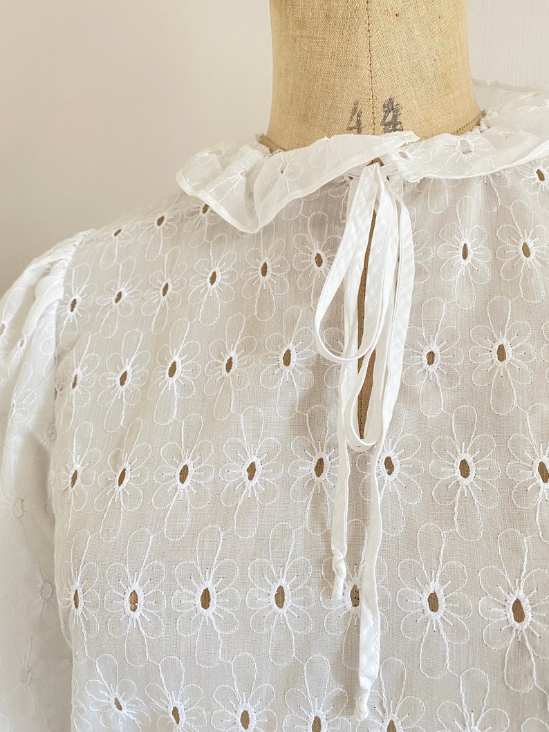 Puede incluir: Blusa blanca de algod&oacute;n con cuello fruncido y un lazo en el cuello. La blusa est&aacute; bordada con un patr&oacute;n floral.