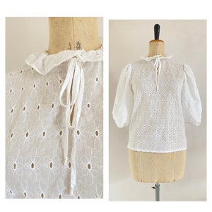 Puede incluir: Blusa blanca de algod&oacute;n con cuello de volantes y un lazo en el cuello. La blusa tiene un patr&oacute;n floral y est&aacute; hecha de una tela ligera.