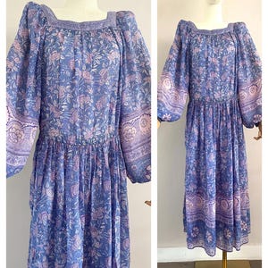 Könnte beinhalten: Ein langes, fließendes Kleid in Blau- und Lavendeltönen mit floralem Muster. Das Kleid hat einen quadratischen Ausschnitt, lange Ärmel und eine geraffte Taille. Die Ärmel und der Saum sind mit einer dekorativen Bordüre verziert.